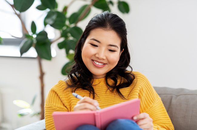 Happy smiling asian young woman in yellow sweater with diary or notebook sitting on sofa at home