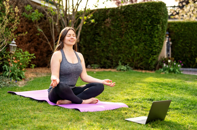 Woman outside on a yoga mat meditating with laptop in front of her