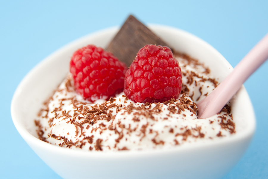 Close up of Greek yogurt topped with raspberries, a dark chocolate square and chocolate shavings