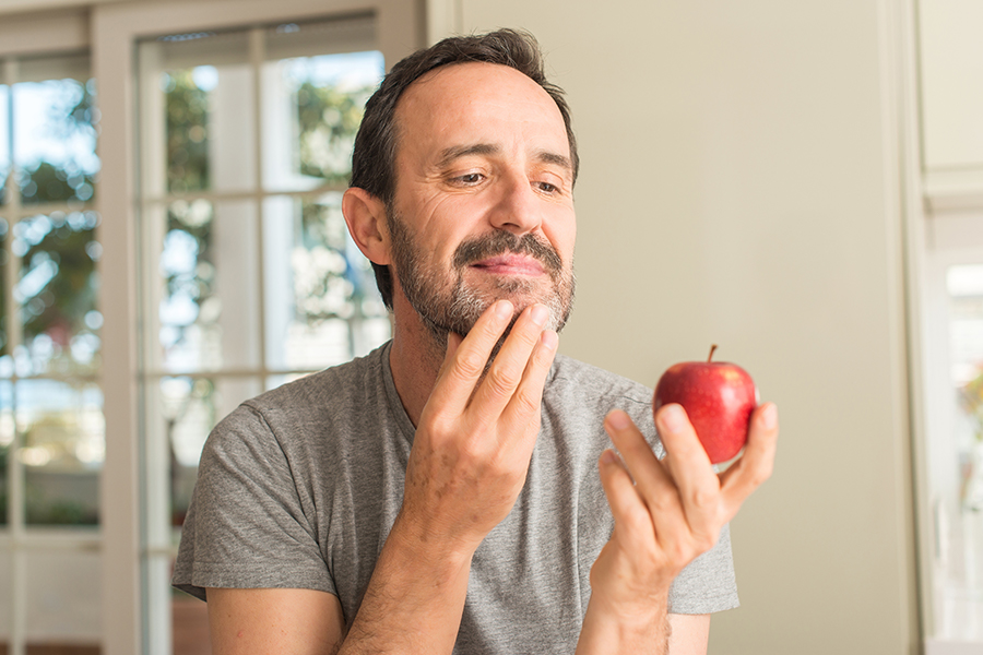 Middle age man holding and looking at red apple with serious face, thinking