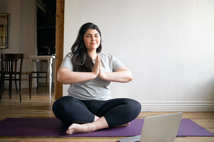 Overweight barefoot young female in sportswear sitting on yoga mat with eyes closed, meditating, having peaceful look, hoding hands in namaste gesture.