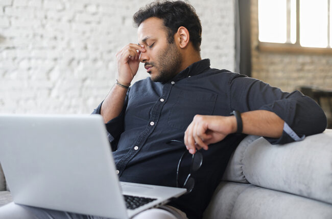 Indian man sitting on couch with laptop, hand pinching nose, tired and stressed