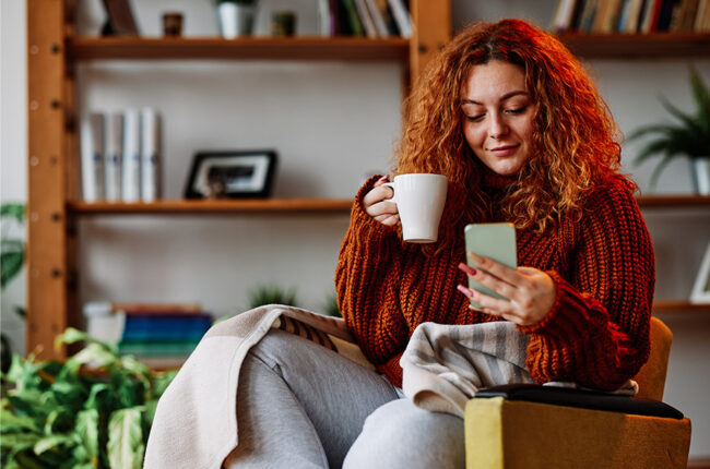 Curly red-headed woman sitting in living room chair sipping tea/coffee scrolling on cellphone