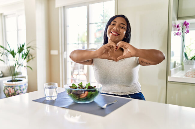 Young hispanic woman eating healthy salad at home smiling in love doing heart symbol shape with hands. romantic concept.