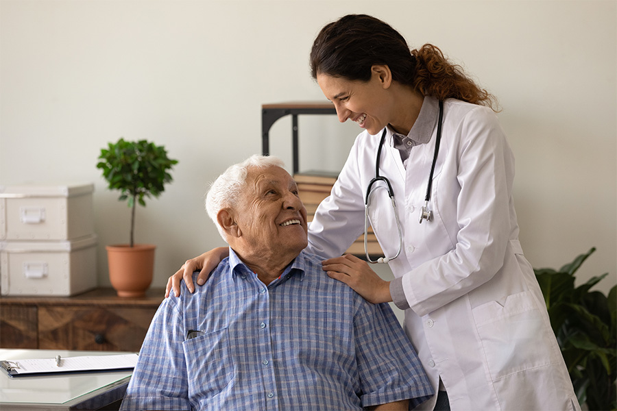 Optimistic young lady doc embrace shoulders of laughing old man sitting on chair at clinic office.