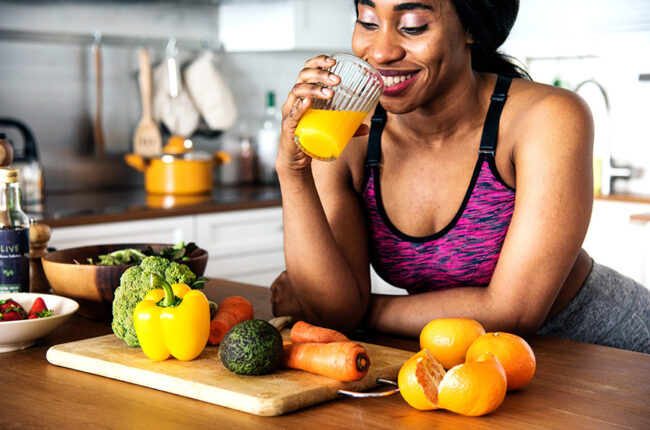 Black woman in exercise clothes, leaning against kitchen counter with vegetables and fruits on a cutting board, drinking orange juice