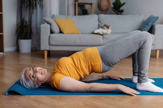 Sporty aged woman doing half bridge yoga pose, strengthening her abs muscles, training in living room, copy space