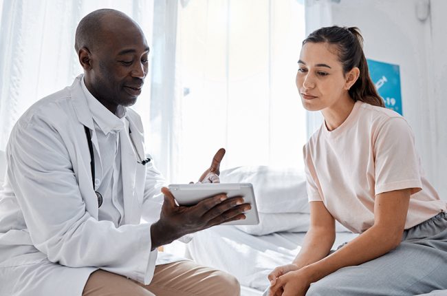 Black doctor with iPad talking to Hispanic female patient in examination room